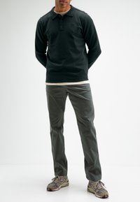 Dunkelgrünes Langarm-Polohemd mit drei Knöpfen, über einem hellen T-Shirt getragen. Olivgrüne Hose und graue Sneakers mit gemusterten Akzenten.