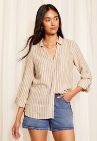 Šviesiai rudos ir baltos spalvų dryžuota marškiniai su sagomis, apykakle ir viena krūtinės kišenė, derinami su džinsiniais šortais. Lygios tekstūros ir atsitiktinio stiliaus.