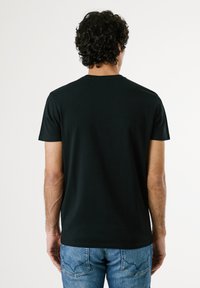 Homme aux cheveux frisés portant un t-shirt noir uni et un jean bleu, debout de dos sur un fond blanc.