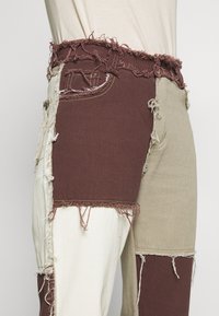 Pantalones de patchwork con secciones de tela marrón, beige y blanca con bordes deshilachados, que muestran una mezcla de texturas y un diseño casual.