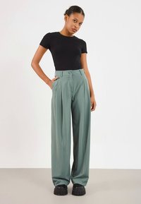 Hooggetailleerde, wijde broekspijpen in zachtgroene stof met voorplooien en een enkele knoopsluiting. Gecombineerd met een aansluitend zwart topje en chunky zwarte schoenen.