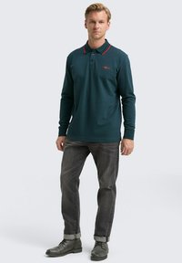 Mann in dunkelgrünem Langarm-Polohemd mit roten Akzenten, hochgekrempelten grauen Jeans und grauen Schnürstiefeln, der vor einem schlichten Hintergrund steht.
