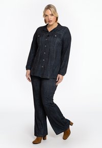 Donkerdenim overhemd en flared broek set met verticale stiksels, knoopsluiting aan de voorkant en lange mouwen. Gecombineerd met tan hoge laarzen.