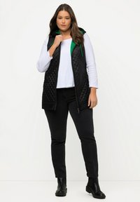 Gepolsterte schwarze Weste mit einer grünen Kapuze, getragen über einem weißen Oberteil, kombiniert mit schwarzen Jeans und Stiefeletten. Verfügt über einen Frontreißverschluss und Taschen.