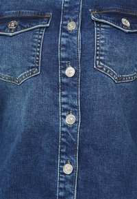 Denim-paita edestä, jossa on metalli-napitus keskiosassa ja kaksi läpällä varustettua rintataskua, yksityiskohtainen tikkaus ja haalistunut pesutekstuuri.