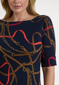 Žena oblečená v tmavě modrém topu se vzorem hnědých pletených pásků, červených popruhů a zlatých řetězů, s rukávy po lokty a kruhovými náušnicemi.