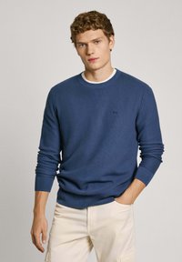 Maglione di cotone blu con finitura testurizzata, scollatura a girocollo e polsini a coste. Logo minimalista sul petto. Indossato con pantaloni color crema.