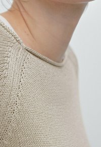 Beige gebreide trui met een ronde halslijn, een textuurpatroon en gedetailleerde stiksels langs de naden. Close-up weergave van het schoudergebied.