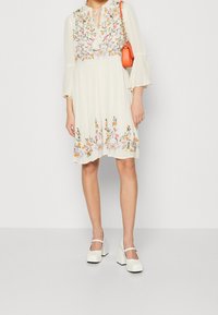 Vit, flödande klänning med blommig broderi i orange, rosa och grönt. Har en knytkrage och utsvängda ärmar. Matchad med vita klackskor med blockhäl.