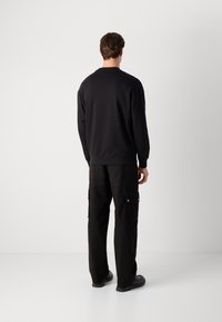 Man standing facing away wearing a black sweatshirt, black cargo pants, and black shoes against a plain white background.