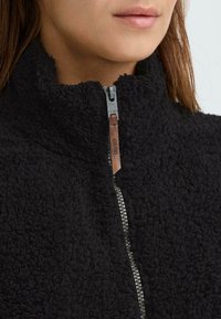 Zwarte fleece jas met een hoge kraag, voorzien van een zilveren rits en een leren trekstrip in bruin, met een gestructureerd, zacht oppervlak.