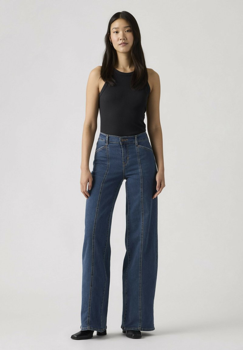 Vrouw staand, gekleed in een zwarte mouwloze top en een high-waisted, wijd uitlopende blauwe jeans met zwarte schoenen, tegen een effen achtergrond.