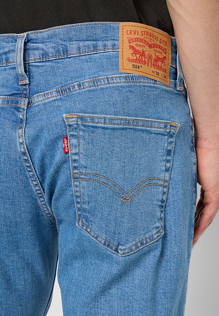 Jeans en denim bleu clair avec une patch en cuir marron à la taille arrière, comportant des coutures contrastantes et un design classique des poches.
