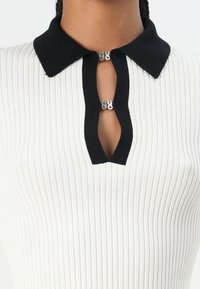White ribbed knit top featuring a black collar and a keyhole opening secured with a silver logo clasp. Texture is smooth and fitted.