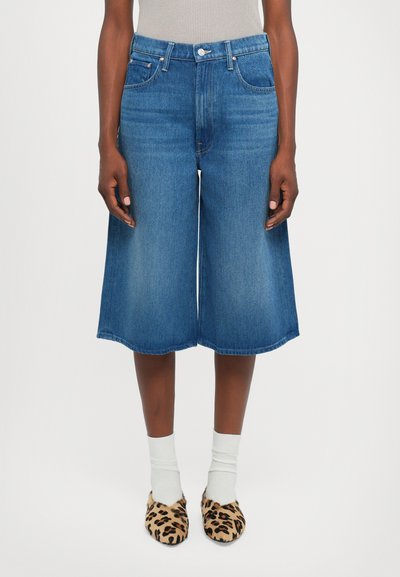Personne portant un short en denim bleu mi-long, des chaussettes blanches et des chaussures à enfiler à imprimé léopard, se tenant devant un fond clair.