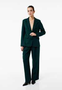 Donkergroene blazer met een diepe V-hals en enkele knoopsluiting, gecombineerd met bijpassende wijde broek en puntige zwarte hakken.