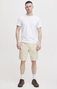 T-shirt blanc à manches courtes associé à un short cargo beige clair avec des poches latérales. Porté avec des chaussures à motifs marron et noir, chaussettes blanches.