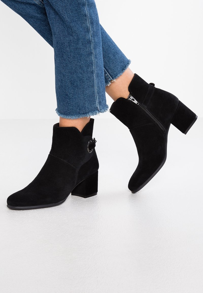 Bottines en suède noir avec un talon carré, une fermeture éclair latérale et un bout arrondi. Elles présentent un accent décoratif en forme de cercle sur le côté extérieur.