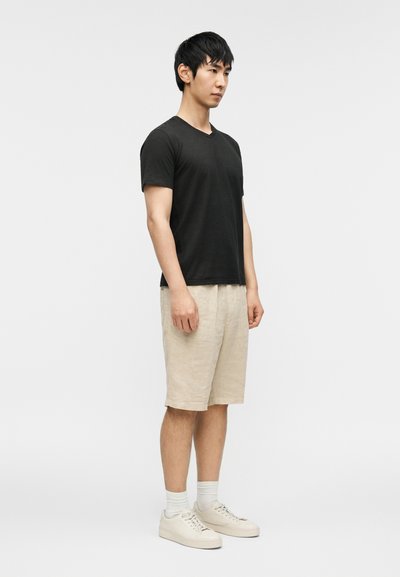 Μαύρο μπλουζάκι με v-neck, μπεζ σορτς λινό, λευκές κάλτσες και ελαφριά αθλητικά παπούτσια. Το ντύσιμο έχει χαλαρή εφαρμογή με ελάχιστες λεπτομέρειες.