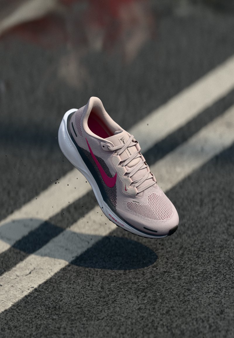Baskets de course Nike rose clair avec un swoosh rose foncé et une semelle blanche, survolant une route en bitume avec des marquages blancs.