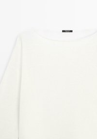 Pull en maille blanche avec un large col rond et une étiquette noire à l'intérieur du col affichant les informations de la marque.