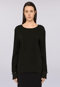 Una donna con i capelli scuri e lisci indossa un top nero a maniche lunghe con scollo tondo e dettagli con lacci sui polsini, in piedi contro uno sfondo neutro.