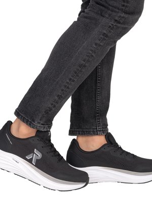 Person trägt schwarze Sportschuhe mit dicken weißen Sohlen und schwarze Jeans, steht mit einem leicht angehobenen Fuß.