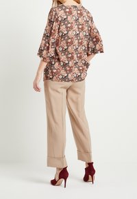 Blouse à motif floral en orange et noir, associée à un pantalon large beige et des chaussures à talons bordeaux. Coupe décontractée avec des manches mi-longues.