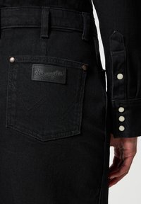 Czarna dżinsowa kurtka z metką Wrangler, z zaokrąglonym designem tylnej kieszeni, metalowymi guzikami i teksturowanym wykończeniem tkaniny.