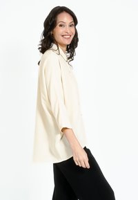 Bēša krekls ar pogām un brīvu piegriezumu, ar trīs ceturtdaļu piedurknēm. Gluda auduma struktūra ar smalku spīdumu, apvienots ar melniem biksēm.