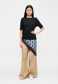 Femme aux longs cheveux foncés portant un haut noir à manches courtes, un pantalon large beige, des chaussures blanches et un panneau de jupe asymétrique à motifs bleus.