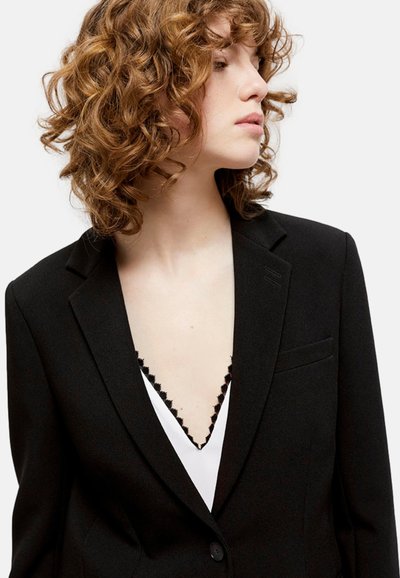 Blazer nero con revers, chiusura a un bottone e tasca frontale. Indossato sopra una parte superiore bianca con bordi scallop in nero.