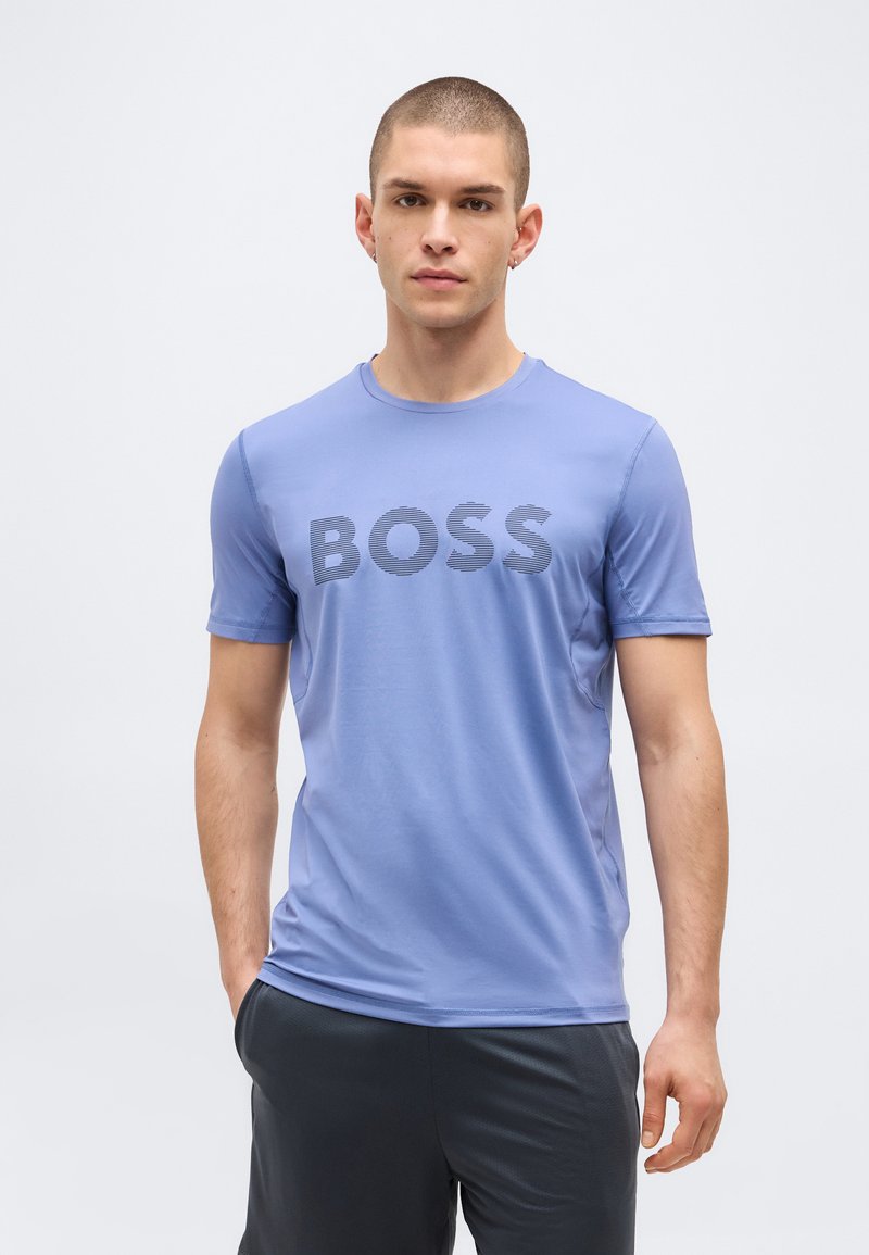 Νεαρός άνδρας με κοντά μαλλιά φορώντας ανοιχτό μπλε μπλουζάκι με το κείμενο «BOSS» και σκουρόχρωμο σορτς, στέκεται απέναντι σε απλό φωτεινό φόντο.