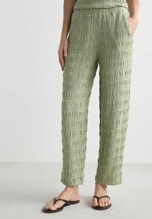 Personne portant un pantalon taille haute texturé vert clair et des tongs noires, debout avec une main dans la poche sur un fond clair.