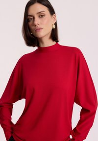 Kobieta z krótkimi ciemnymi włosami, ubrana w luźną czerwoną bluzkę z długim rękawem i złotymi kolczykami w kształcie kół, na jasnym tle.