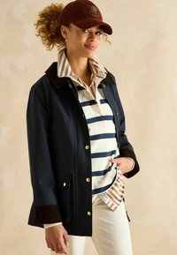 Jachetă navală, impermeabilă, cu butoane aurii și manșete din căptușeală maro, purtată peste o cămașă cu dungi în nuanțe de bej și bleumarin.