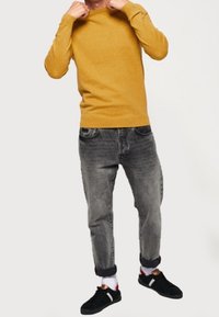 Maglione giallo senape a maniche lunghe abbinato a jeans grigi scoloriti. Sneakers nere con dettagli bianchi. Polsini arrotolati sui jeans.