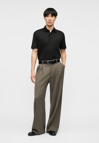 Άνδρας που φοράει μαύρο πόλο με κοντό μανίκι, καφέ παντελόνι με φαρδύ πόδι και μαύρη ζώνη, και μαύρα παπούτσια, στέκεται με το ένα χέρι στην τσέπη.