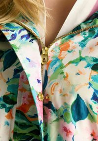 Jas met bloemenpatroon in levendige kleuren en gouden ritsen met een ronde trekker; gladde, lichte stof met een glanzende afwerking.