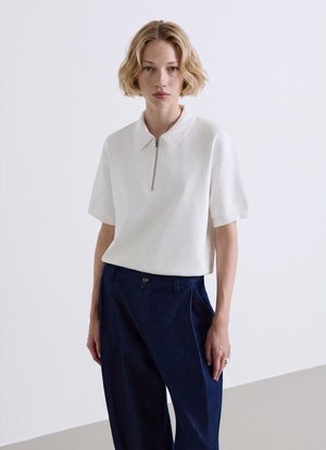 Giovane donna con capelli corti biondi che indossa un top bianco a maniche corte con cerniera e pantaloni a vita alta blu scuro, in piedi su uno sfondo semplice.