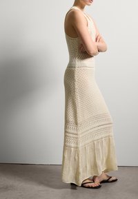 Maxikleid in Creme mit Lochmuster, tailliertem Oberteil und ausgestellt geschnittenem Saum. Kombiniert mit flachen schwarzen Sandalen. Glatte, atmungsaktive Textur.