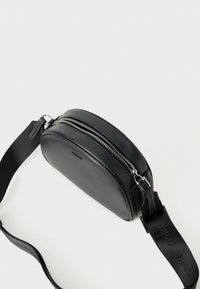 Čierna kožená crossbody taška so zaobleným tvarom, strieborným zipsom a nastaviteľným popruhom so slepým brandingom. Hladká textura, minimalistický dizajn.