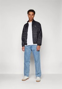 Svart vattert lett jakke med elastiske mansjetter, båret over en hvit t-skjorte og lyseblå jeans, kombinert med hvite joggesko.