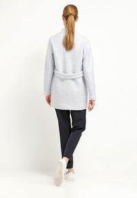 Cappotto grigio chiaro in morbido tessuto, con vita cinturata, maniche lunghe e silhouette dritta. Abbinato a pantaloni neri e sneakers bianche.