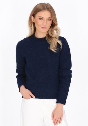 Kobieta z blond włosami, ubrana w granatowy, puszysty sweter i białe spodnie, stoi z jedną ręką w kieszeni na białym tle.