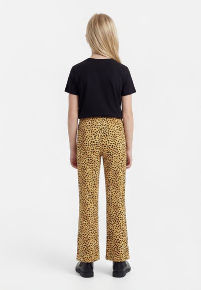 Hosszú szőke hajú gyermek áll háttal, fekete rövid ujjú pólót, sárga leopárdmintás nadrágot és fekete cipőt visel.