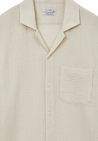 Chemise beige clair coupe décontractée avec col camp, boutonnée devant et une poche poitrine unique en tissu texturé.