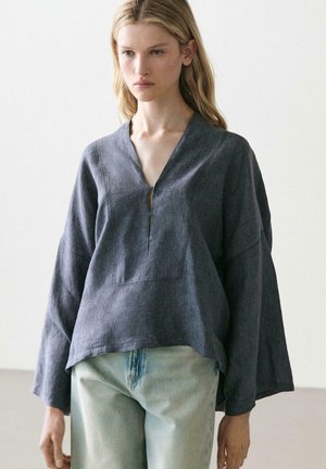 Frau trägt eine locker sitzende dunkelgraue Bluse mit langen Ärmeln und V-Ausschnitt sowie hellblaue hoch taillierte Jeans vor einem einfarbigen Hintergrund.