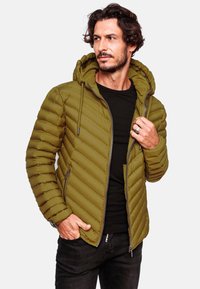 Olijfgroene pufferjack met horizontale stiksels, ritssluiting, zijzakken en verstelbare capuchon. G gedragen over een zwart shirt, met textuur.