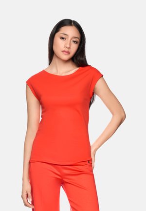 Frau mit langen dunklen Haaren, die ein leuchtend rotes kurzärmeliges Oberteil und passende Hose trägt, steht mit einer Hand in der Hüfte.